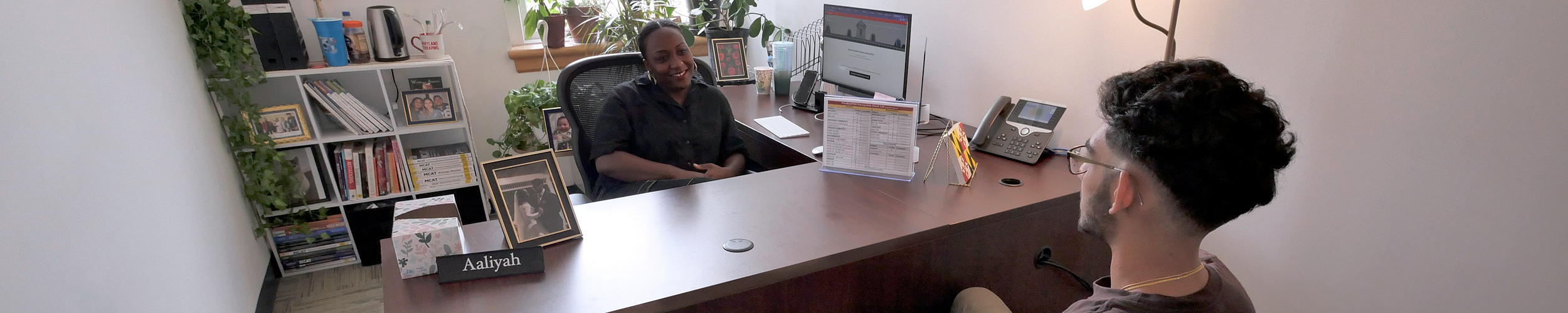 A woman advising a young man in her sunlit office. In the background there is a window, houseplants, and a bookcase containing volumes on the MCAT and health professions.